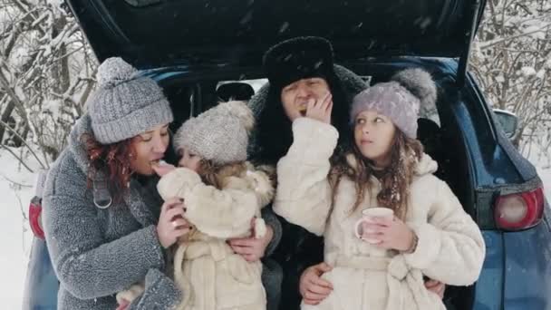 Zimní čajový piknik. Šťastná rodina si dává chutnou svačinku, čajový dýchánek se smetanou, venku. Sedí na kufru auta, v zasněženém lese, během sněžení. zimní rodinná zábava. šťastný čas na zasněžené zimě — Stock video
