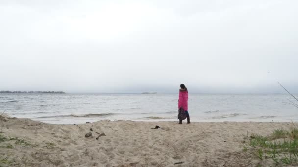 Прогулянка біля моря взимку. Сумно, одна жінка мандрівник спостерігає на морських або річкових хвилях, ходить піщаним пляжем, на вітряний, холодний, похмурий зимовий день. Вид ззаду. прогулянка вздовж пляжу взимку — стокове відео