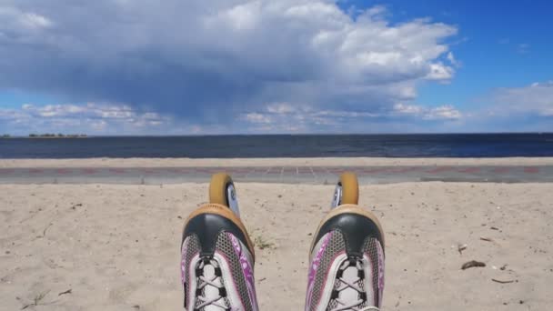 屋外でローラースケート。屋外でのローラーブレード。接近中だ。ローラースケートの足は、砂のビーチ、海、雲のある青空を背景に、ローラーブーツに隠されています. — ストック動画