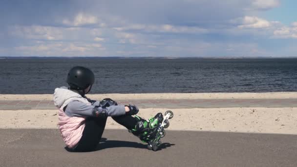 孩子们在打滚。无忧无虑的少女，戴着滚筒和头盔，坐在沙滩旁边的人行道上，在滑旱冰之后休息，在阳光灿烂的春天欣赏着海景。周末愉快 — 图库视频影像