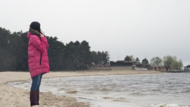 Прогулянка біля моря взимку. Сумна, одна жінка спостерігає на морських або річкових хвилях, гуляє вздовж піщаного пляжу, на сніжному, вітряному, холодному, зимовому дні. прогулянка вздовж пляжу взимку — стокове відео