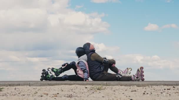 Bruslení venku. Bezstarostný otec a dospívající dívka, v kolečkách sedí na chodníku vedle písečné pláže, odpočívá po kolečkových bruslích, obdivuje mořský plášť na slunném jarním dni. Aktivní víkend — Stock video
