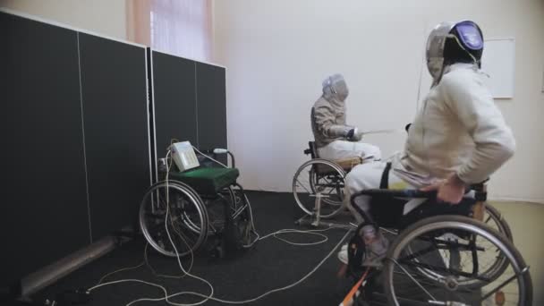 Engelli atlet. Eskrim. Tekerlekli sandalyedeki beyaz eskrim üniformalı eskrimciler, eskrim kasklarıyla yüzlerinde rapier ve idman antrenmanları tutuyorlar.. — Stok video