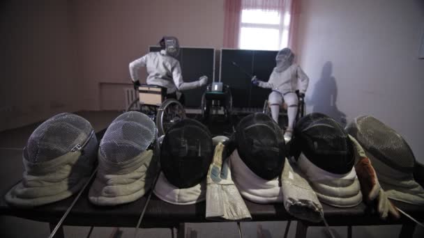 Atleta discapacitado. Esgrima. equipo para la esgrima, cascos, máscaras y guantes están en el telón de fondo del duelo de esgrima sparring de dos atletas de esgrima en silla de ruedas con habilidades físicas reducidas. Practican — Vídeos de Stock