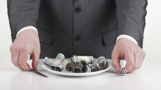 Hombre de negocios y dinero, efectivo. Primer plano. un hombre de negocios, en traje formal de negocios, mezcla billetes de cien dólares enrollados en un plato con cubiertos, cuchillo y tenedor en sus manos. Parece que va a comer. — Vídeos de Stock