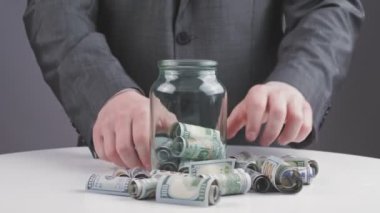 İş adamı, para, para. Yakın plan. İş adamı, resmi takım elbiseli, yüz dolarlık banknotları cam kavanoza koyar. Tasarruf ve mevduat. Finans ve yatırım, başarı kavramı.