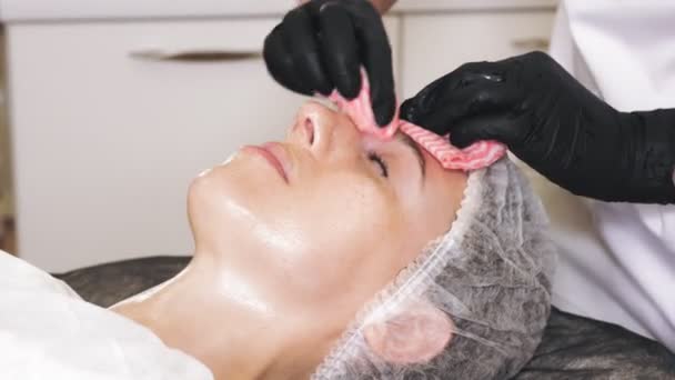 Terapia de beleza. Cosmetologia cuidados com a pele. close-up. cosmetologist, em luvas médicas pretas, retira restos do produto de cara cosmético, da cara feminina com toalhetes molhados especiais. procedimento anti envelhecimento. Facial — Vídeo de Stock