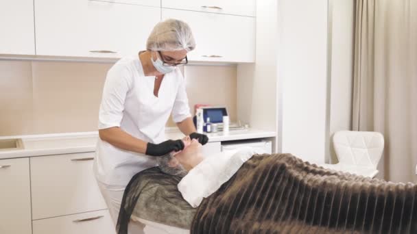 Beauty therapy. Cosmetology skin care. cosmetologist, in medical mask and gloves, removes remains of cosmetic skincare facial scrub from female face with special wet wipes, at beauty clinic. anti — Stockvideo