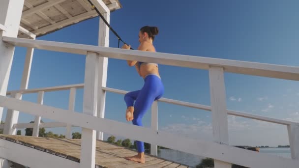 屋外でワークアウト。サスペンションストラップ。ストラップ付きワークアウト。陸上の若い女性は、日没または日の出の間にビーチでループTRXを使用して全身抵抗運動をしています。フィットネストレーニング — ストック動画