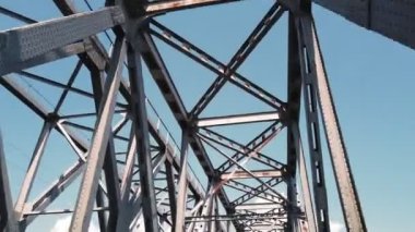 Metal köprü. Mavi gökyüzüne karşı kaba demir yapılar, hareket halindeki bir arabadan çekiliyor. Alt manzara. Nehrin karşısındaki demiryolu ve yol köprüsü. Çelik kirişleri aşan siluet.