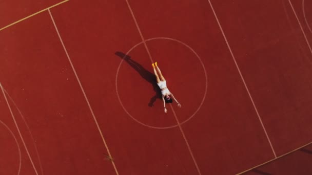 Fitness buiten. Aero, bovenaanzicht. Sportieve vrouw in witte top en korte broek, doet verschillende oefeningen. achtergrond van oranje hof met witte markeringen. Buitensporten. Zomer dag. in het stadion — Stockvideo