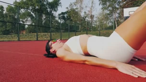 Fitness all'aperto. Donna sportiva, in top bianco e pantaloncini, sneakers arancioni e occhiali da sole, sta facendo vari esercizi su campo arancione con marcature bianche, allo stadio. giorno d'estate. Sport all'aria aperta — Video Stock