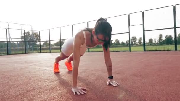 Fitness à l'extérieur. Femme sportive, en haut blanc et short, baskets orange et lunettes de soleil, fait divers exercices sur terrain orange avec des marques blanches, au stade. jour d'été. Sports de plein air — Video