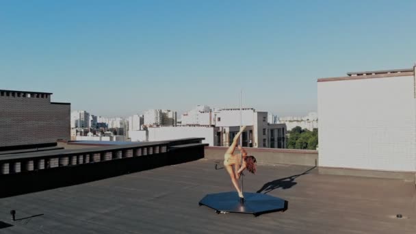 Paaldansen. Luchtvaart. fit vrouwelijke turnster, in beige maillot, voert acrobatische oefeningen op metalen roterende paal, op wolkenkrabber dak, met stedelijk uitzicht op de stad. ochtend boven de stad. — Stockvideo