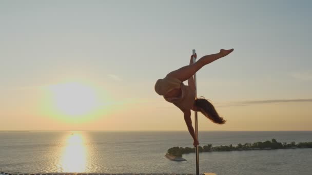 Direk dansı. Silüet. uygun kadın jimnastikçi, bej taytlı, akrobatik egzersizler yapıyor metal döner direkte, gökdelen çatısında, deniz manzaralı arka planda — Stok video