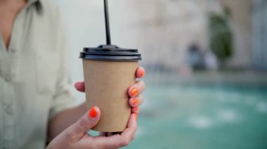 Paket kahve. Kahve. Sütlü. Kahve içeceğim. Yakın plan. Kadın elleri, şehir çeşmesinin arka planında plastik kapaklı ve pipetli tek kullanımlık kahverengi bir kağıt bardağı tutuyor.