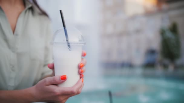 牛奶鸡尾酒 喝酒去了 把它带走 在城市喷泉的背景下 女性的手拿着奶昔 放在透明的一次性塑料杯中 配有塑料盖和吸管 — 图库视频影像