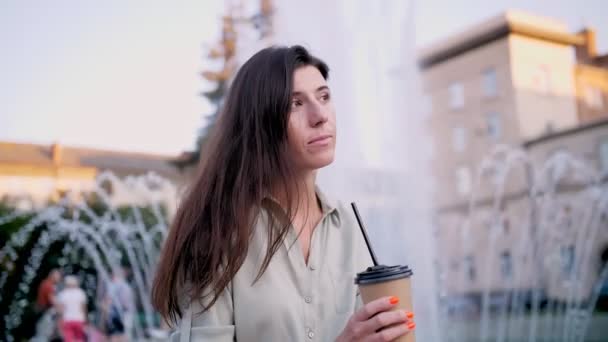 Caffè da portare via. caffè. latte macchiato. bevanda al caffè. una giovane donna beve attraverso una cannuccia il suo latte da un bicchiere di carta usa e getta, seduta accanto a una fontana, in una sera d'estate. — Video Stock