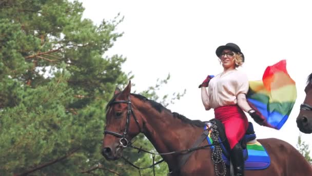 Lgbt Regenbogenfahne. Reiten. schönes Reitermädchen, mit flatternder Regenbogenfahne in den Händen posiert beim Reiten im Wald — Stockvideo
