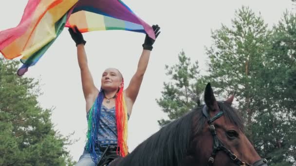 Szivárvány zászló. Lovas lovaglás. gyönyörű lovas lány, szivárvány haj raszta és csapkodó szél lgbt szivárvány zászló, pózol, miközben lovaglás, az erdőben — Stock videók