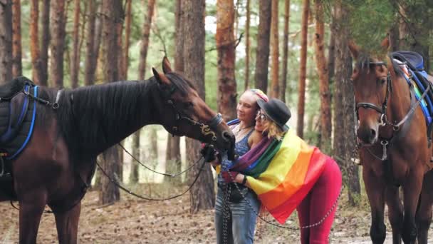 Lgbt. Regenbogenfahne. Gleichgeschlechtliche Liebe. Das junge lesbische gleichgeschlechtliche Paar betreibt ein gemeinsames Hobby, das Reiten. Frauen umarmen sich, haben Spaß, neben ihren Pferden, im Wald — Stockvideo