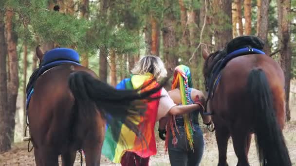 Lgbt. Regenbogenfahne. Das junge lesbische gleichgeschlechtliche Paar betreibt ein gemeinsames Hobby, das Reiten. Händchenhaltend gehen Frauen durch den Wald, neben ihren Pferden. Rückseite. Gleichgeschlechtliche Liebe. — Stockvideo