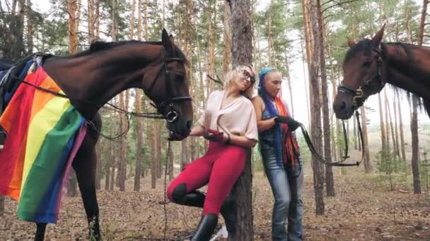 Lgbt. bandiera arcobaleno. giovane lesbica coppia dello stesso sesso è impegnata in hobby comune, equitazione. le donne stanno trattando i loro cavalli con chicche, mentre riposano durante le passeggiate nella foresta. amore dello stesso sesso. — Video Stock