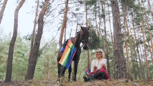 Lgbt bandera del arco iris. equitación. hermosa chica jinete, con la bandera del arco iris lgbt, posando junto a su caballo, mientras está sentado y descansando durante la caminata en el bosque. — Vídeos de Stock