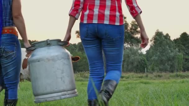 Leche de vaca fresca. Dos mujeres agricultoras llevan juntas, una lata grande con leche fresca a través del prado verde, al atardecer, contra el fondo de la vaca pastando. ordeño. granja lechera. Productos lácteos. Agricultura. — Vídeos de Stock