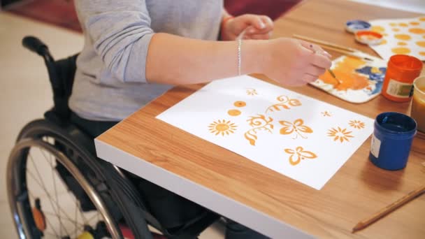 Αμαξίδιο. αναπηρία. Κοντινό πλάνο. ανάπηρη νεαρή γυναίκα ζωγραφίζει, κάθεται σε αναπηρικό καροτσάκι στο τραπέζι. δημιουργικά μαθήματα για άτομα με αναπηρίες. Εκπαίδευση, σχέση και ανθρωπιά — Αρχείο Βίντεο
