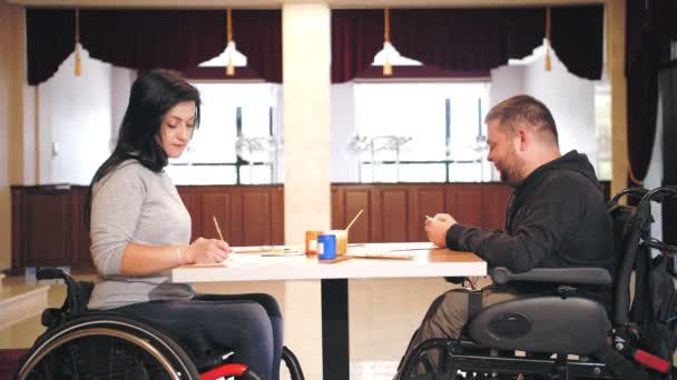 Інвалідне крісло. інвалідність. неповносправний молодий чоловік і жінка малюють фарбами, сидячи в інвалідних візках за столом. Уроки творчої групи для людей з обмеженими можливостями. Освіта, відносини та людяність — стокове відео