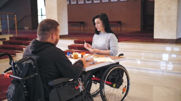 Cadeira de rodas. deficiência. homem e mulher jovens deficientes estão desenhando com tintas, sentados em cadeiras de rodas à mesa. aulas de grupo criativo para pessoas com deficiência. Educação, relacionamento e humanidade — Vídeo de Stock