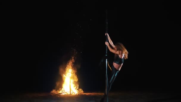 Pole dance. Hexenhalloween. Fitte Turnerin, in schwarzem Lederoutfit und High Heels, führt akrobatische Übungen auf einer rotierenden Stange aus Metall aus, nachts, im Licht des großen Feuers im Hintergrund. — Stockvideo