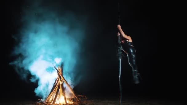 Baile pole. Bruja Halloween. gimnasta femenina en forma, en traje de cuero negro y tacones altos, realiza ejercicios acrobáticos en poste giratorio de metal, por la noche, a la luz de un gran fuego en el fondo. — Vídeo de stock