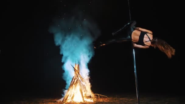 Pole dance. Halloween vrăjitoare. se potrivesc gimnast feminin, în costum din piele neagră și tocuri înalte, efectuează exerciții acrobatice pe pol rotativ metalic, pe timp de noapte, prin lumina focului mare în fundal . — Videoclip de stoc