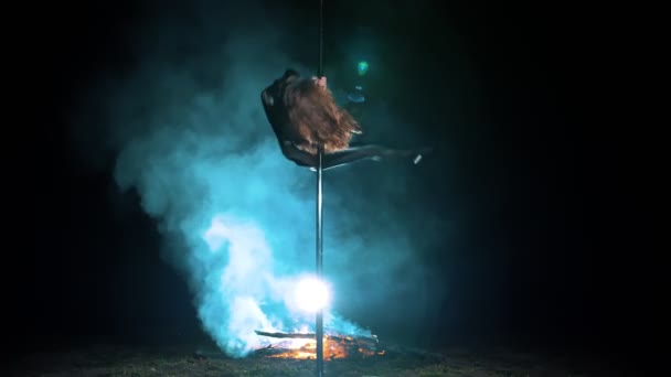 Baile pole. Bruja Halloween. gimnasta femenina en forma, en traje de cuero negro y tacones altos, realiza ejercicios acrobáticos en poste giratorio de metal, por la noche, a la luz de un gran fuego en el fondo. — Vídeo de stock