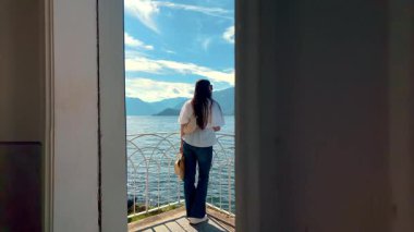Beyazlı kadın Como Gölü manzaralı panoramik pencerenin yanında duruyor. Teras girişi olan lüks bir iç mekan..
