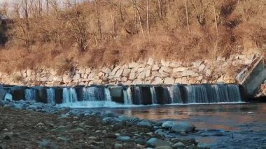 Nehirdeki küçük beton eşiği ya da baraj yapay şelale oluşturuyor. Yumuşak güneş ışığı altında arka planda kış çalılıkları.