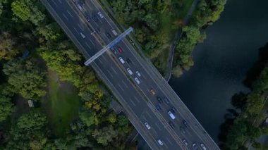 Hava görüntüsü, ağaçlarla çevrili bir nehrin üzerinden geçen yoğun bir karayolu trafiğini gösteriyor. İHA kamerası, rotasyonu sol İHA 'ya yöneltiyor 