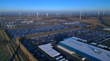 Depoları ve tren rayları olan, rüzgar türbinleriyle çevrili büyük bir dağıtım merkezinde yeni arabalar bekliyor. panorama genel görünüm dronu