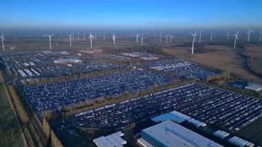 Depoları ve tren rayları olan, rüzgar türbinleriyle çevrili büyük bir dağıtım merkezinde yeni arabalar bekliyor. İHA alçalıyor