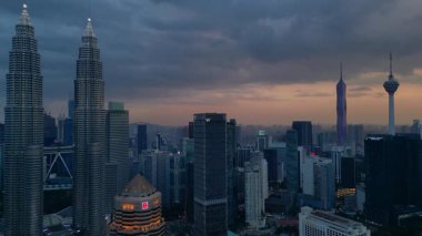 Kuala Lumpur şehrinin silueti, kasvetli bir günbatımı sırasında diğer modern gökdelenlerle çevrili ikonik Petronas İkiz Kuleleri ile çevrilidir. panorama genel görünüm dronu