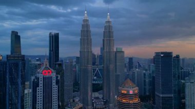 Kuala Lumpur şehrinin silueti, kasvetli bir günbatımı sırasında diğer modern gökdelenlerle çevrili ikonik Petronas İkiz Kuleleri ile çevrilidir. Ters İHA uçuşu