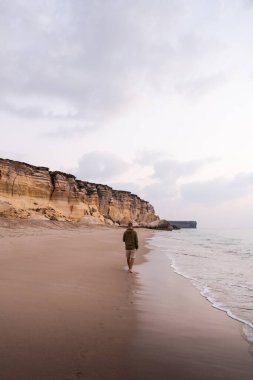Okyanus dalgaları ve yüksek uçurumların yanında kumsalda yalınayak yürüyen bir adam.