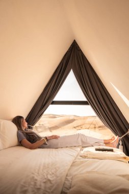 Lüks bir çadırda yatakta dinlenen bir kadın çöl manzarasına bakıyor.