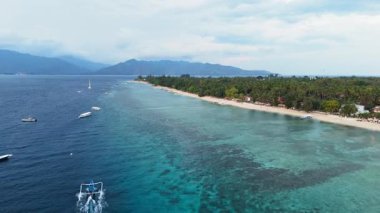 Kumlu bir sahil, turkuaz bir okyanus ve yakınlarda yüzen tekneler olan tropikal bir adanın havadan görünüşü.