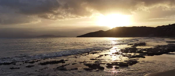 Kara bulutlar ve kayalık sahile yansıyan altın ışıkla denizin üzerinde dramatik bir gün doğumu. Güçlü kontrast ve güçlü kıyı atmosferi.