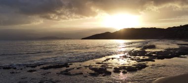 Kara bulutlar ve kayalık sahile yansıyan altın ışıkla denizin üzerinde dramatik bir gün doğumu. Güçlü kontrast ve güçlü kıyı atmosferi.