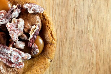 Close-up of a caramel cookie topped with glazed pecan and sugared nut pieces on a wooden surface.