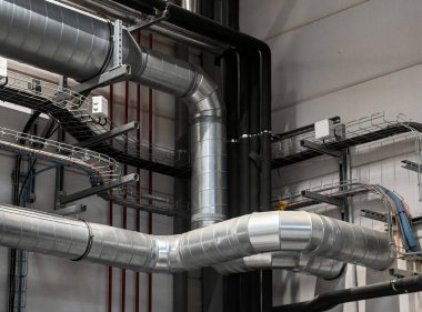 Industrial metal ductwork with bends and joints, part of HVAC system, mounted near ceiling with pipes and cable tray in background.